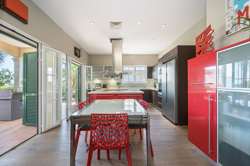 Cuisine lumineuse avec îlot rouge, électroménager en inox et table à manger près des portes vitrées.