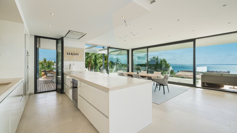 Cuisine blanche moderne avec grand îlot, salle à manger et baies vitrées du sol au plafond donnant sur l'océan.