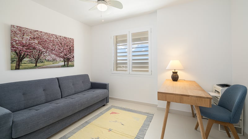 Pièce minimaliste avec un canapé gris, un bureau en bois avec une lampe, une chaise bleue et une fenêtre avec volets blancs.