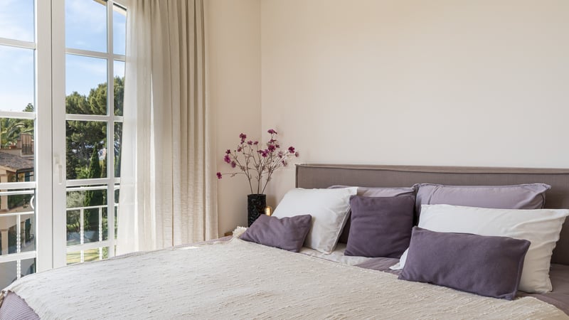 Une chambre lumineuse avec un lit aux coussins mauves à côté d'une grande fenêtre donnant sur des pins verdoyants.