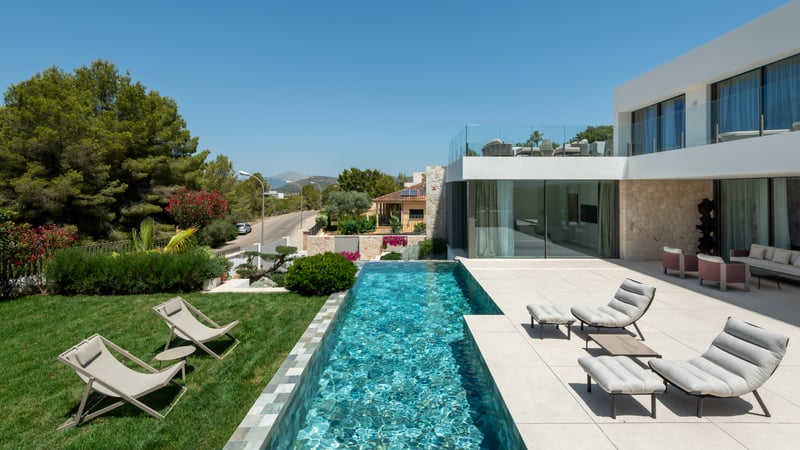 Villa de luxe blanche avec piscine à débordement, chaises longues sur terrasse en pierre et pelouse verte.