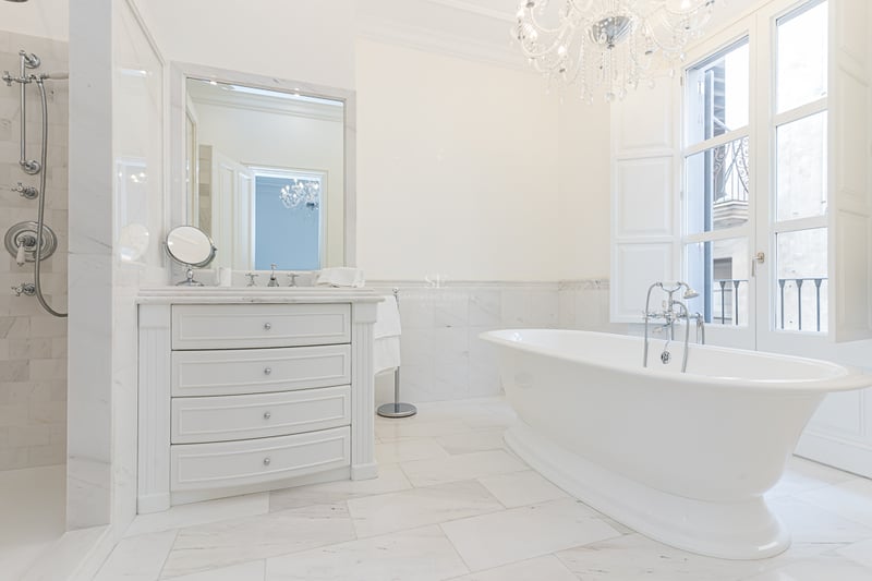 Salle de bain de luxe en marbre blanc avec baignoire îlot, lustre en cristal et grande fenêtre lumineuse.