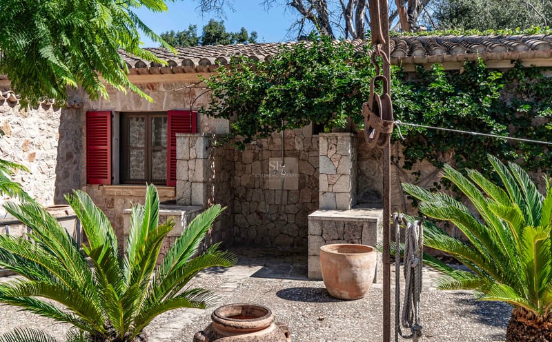Espace de douche extérieure en pierre avec volets rouges, pots en terre cuite et plantes vertes dans une cour méditerranéenne.