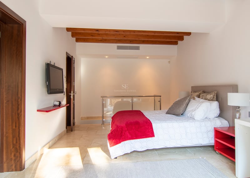 Chambre moderne avec lit double, touches de rouge, poutres en bois au plafond et sol en pierre crème.