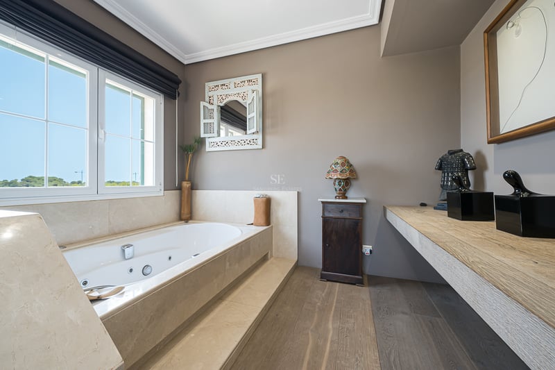 Salle de bain ensoleillée avec baignoire balnéo en marbre, parquet en bois et vue extérieure.