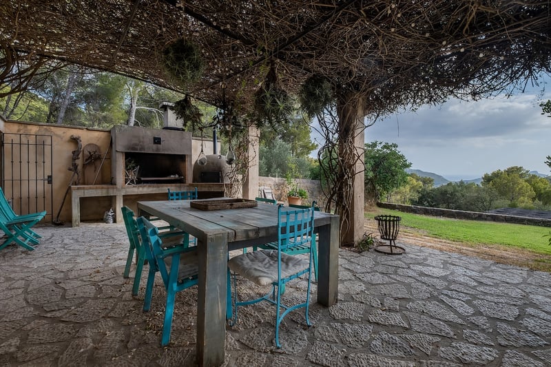 Terrasse en pierre rustique avec chaises turquoise, table en bois et barbecue sous une pergola de vigne.