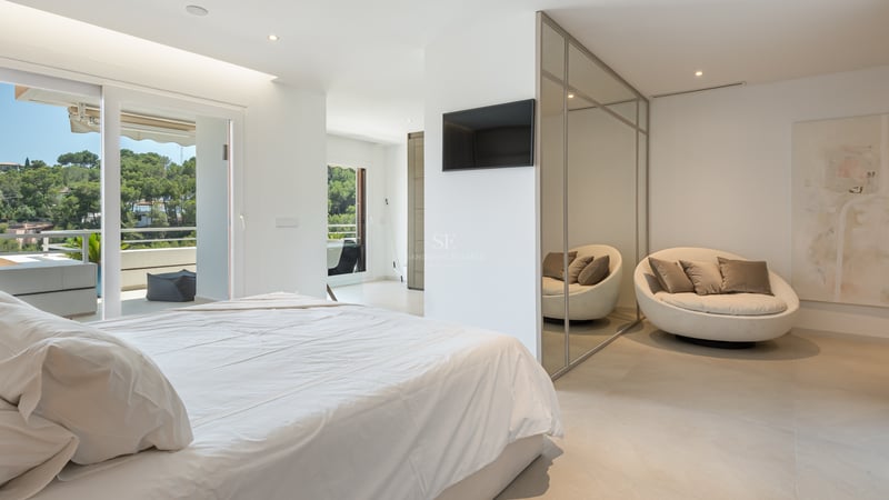 Chambre lumineuse avec lit blanc, armoire en miroir, fauteuil circulaire et vue sur la terrasse et les arbres.