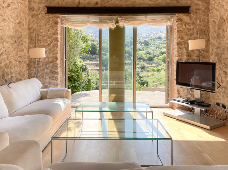 Salon lumineux doté de murs en pierre naturelle, canapés blancs et baies vitrées ouvrant sur des collines verdoyantes.