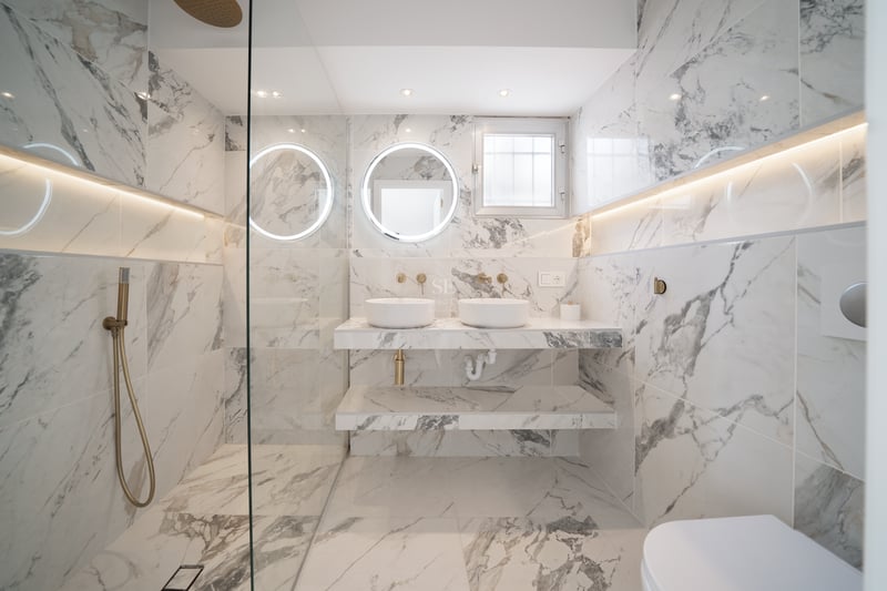Salle de bains de luxe avec murs en marbre, double vasque, miroirs circulaires rétroéclairés et robinetterie en laiton.