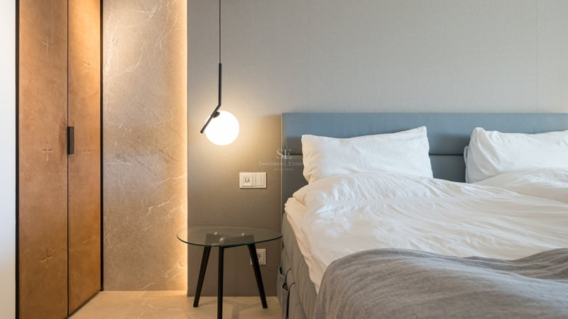 Chambre luxueuse avec tête de lit bleue, table en verre, suspension moderne et mur en pierre éclairé.