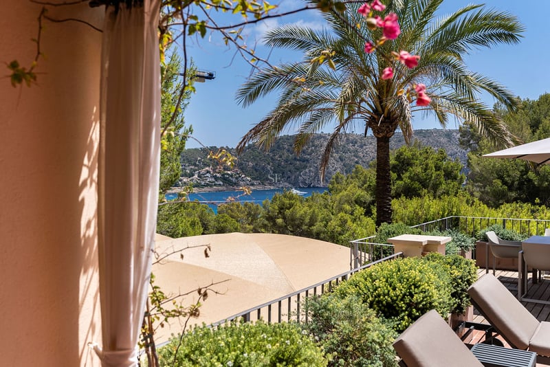 Terrasse avec chaises longues et coin repas offrant une vue sur une baie bleue et des montagnes verdoyantes.