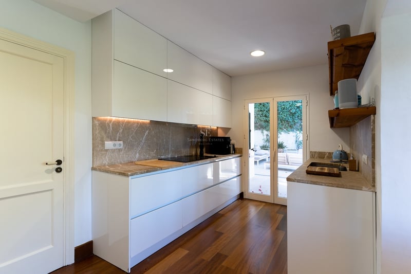 Cuisine moderne blanche avec plans de travail en marbre, parquet et portes vitrées ouvrant sur une terrasse.