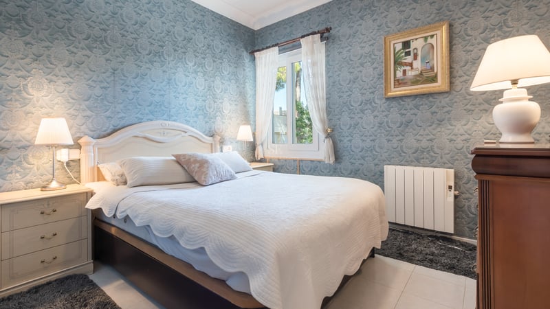 Chambre de style classique avec papier peint bleu à motifs, lit blanc et tables de chevet en bois sous une lumière douce.