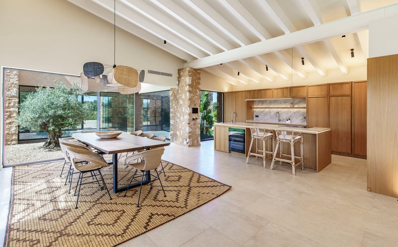 Cocina abierta con isla de madera, comedor contiguo, vigas blancas y pared de piedra natural.