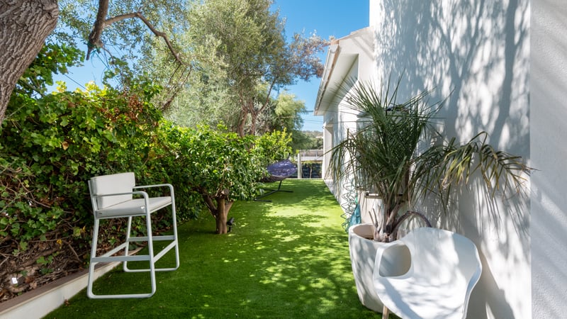 Petit jardin latéral avec gazon synthétique, chaises design blanches, arbres verdoyants et mur de villa blanc.