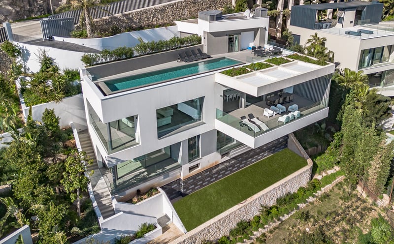 Vue aérienne d'une villa blanche à trois niveaux avec piscine à débordement sur le toit et balcons en verre.