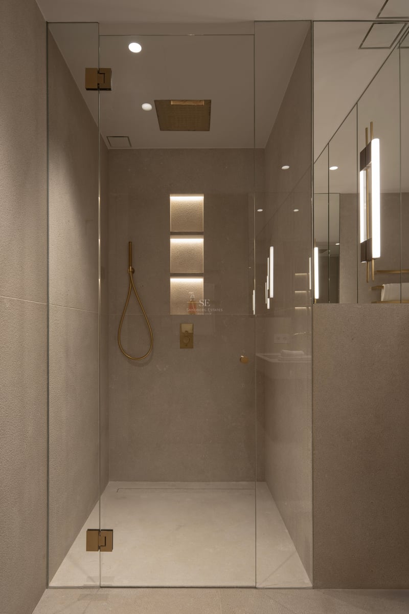 Douche en verre sans cadre avec carreaux de pierre couleur sable, éclairage encastré et robinetterie en or brossé.