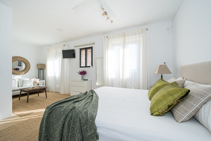 Chambre principale lumineuse avec un grand lit, coussins verts, un coin salon avec canapé blanc et tapis en jute.