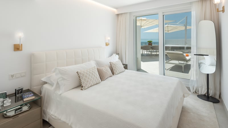 Chambre principale lumineuse avec tête de lit capitonnée et baies vitrées ouvrant sur une terrasse face à la mer.