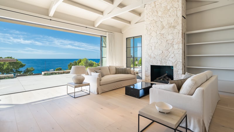 Salon lumineux avec cheminée en pierre, canapés crème et baies vitrées ouvrant sur la Méditerranée.