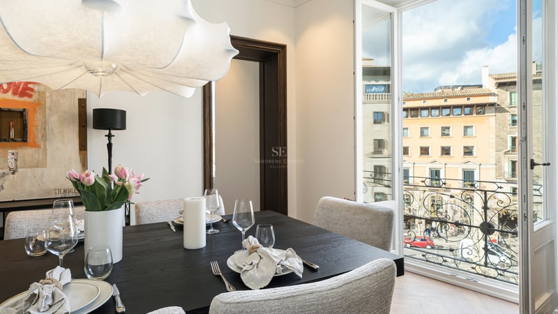 Modern dark wood dining table and white chairs facing an open balcony overlooking historic city architecture.