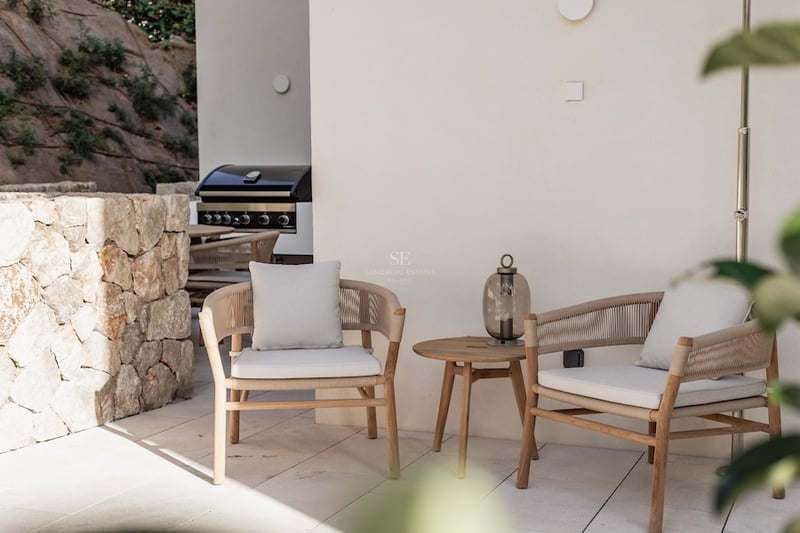 Deux élégants fauteuils modernes avec coussins à côté d'une table en bois sur une terrasse en pierre avec un barbecue.