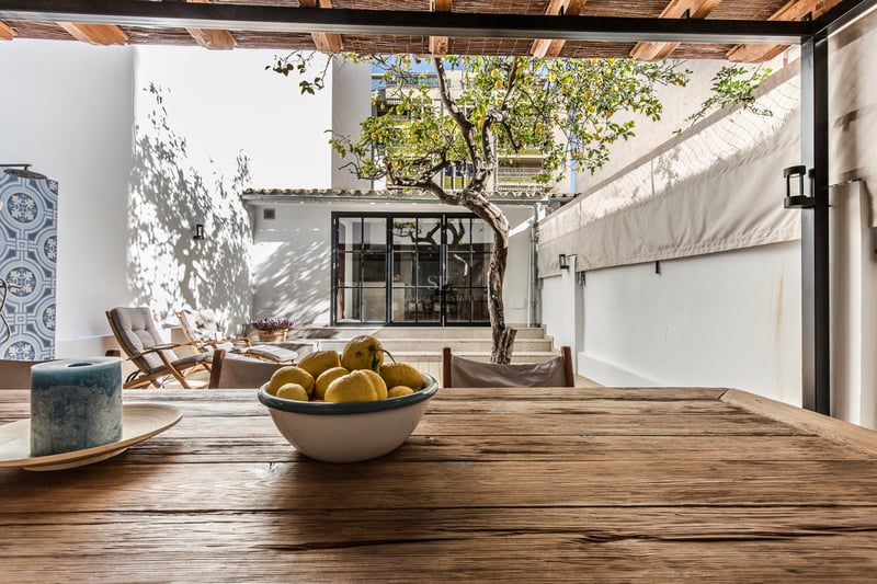 Terrasse ensoleillée avec table en bois, citronnier, chaises longues et murs blancs sous un toit en roseau.