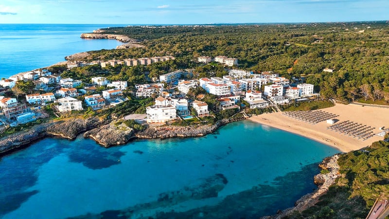 Drönarvy över en turkos vik med sandstrand omgiven av vita byggnader och lummig grön skog.