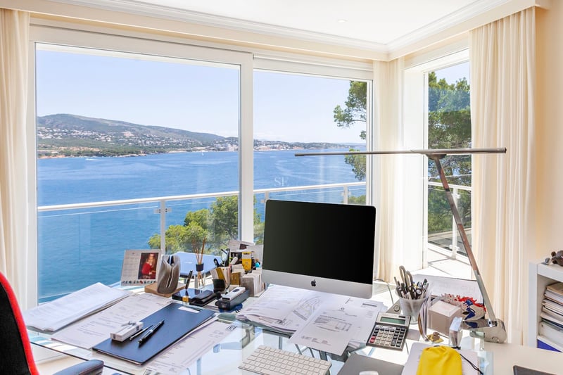 Bureau moderne avec table en verre et ordinateur devant une vue imprenable sur la mer bleue et la côte méditerranéenne.