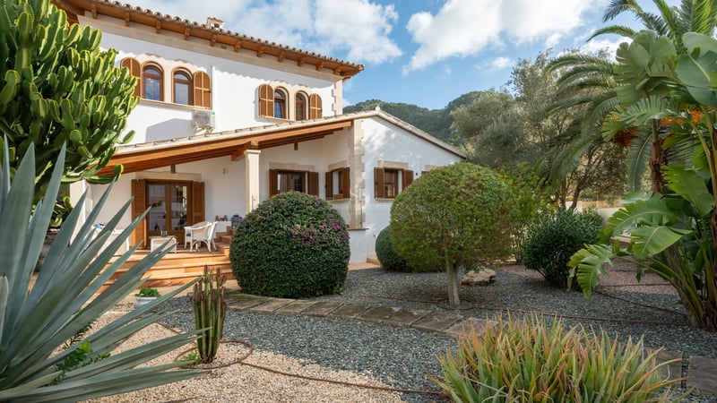Villa majorquine blanche avec volets en bois et jardin de cactus sur fond de montagnes.