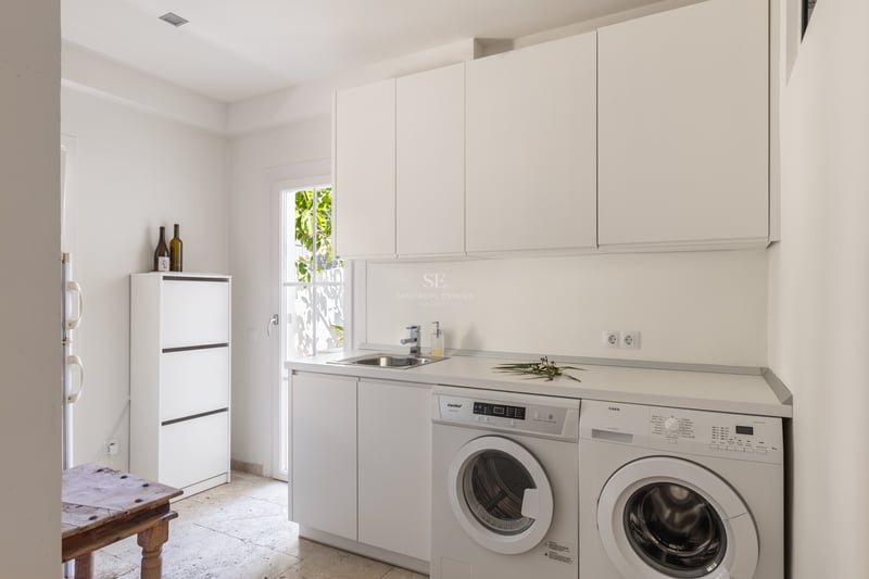 Buanderie blanche avec lave-linge, sèche-linge et évier en inox sur un sol en pierre naturelle claire.