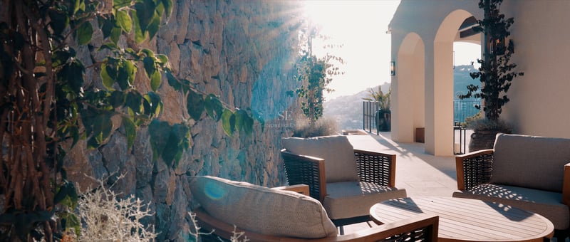 Terrasse ensoleillée avec fauteuils gris élégants, table en bois, mur en pierre brute et piliers blancs arqués.