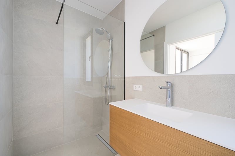 Salle de bain moderne avec douche vitrée, carrelage gris, miroir circulaire et meuble vasque en bois.