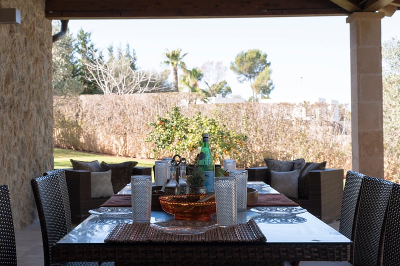 Terrasse méditerranéenne couverte avec salle à manger en osier, murs en pierre naturelle et vue sur un jardin verdoyant.