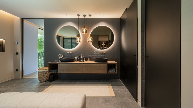 Salle de bain moderne avec deux miroirs ronds éclairés, vasques noires et meuble en bois sur sol en pierre.