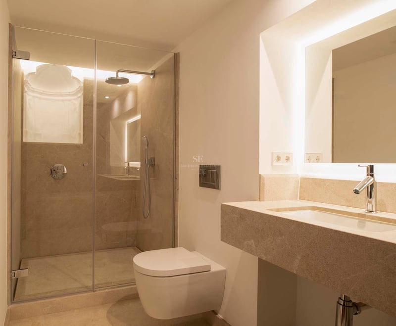 Salle de bain moderne avec douche vitrée, toilettes suspendues et vasque en pierre avec miroir rétroéclairé.