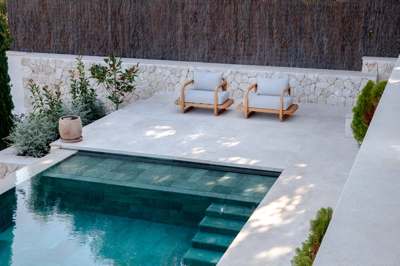 Vue de dessus d'une piscine moderne aux carreaux turquoise à côté d'une terrasse en pierre avec deux fauteuils en bois.