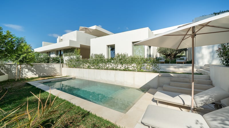 Villa blanche moderne avec piscine rectangulaire, patio en pierre, bains de soleil et jardin verdoyant sous un ciel bleu.