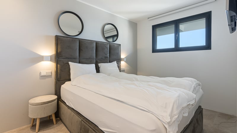 Dormitorio moderno con cabecero de terciopelo gris, espejos circulares, apliques de pared y ventana de aluminio negro.