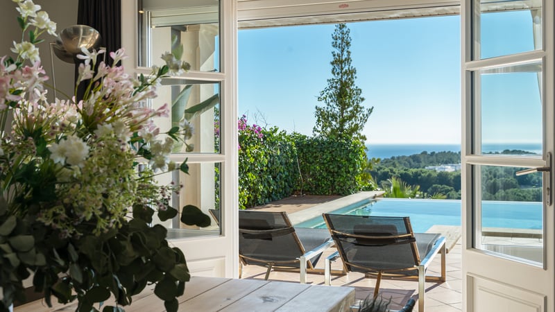 Vue à travers des portes-fenêtres ouvertes sur une terrasse ensoleillée avec chaises longues, piscine et mer.