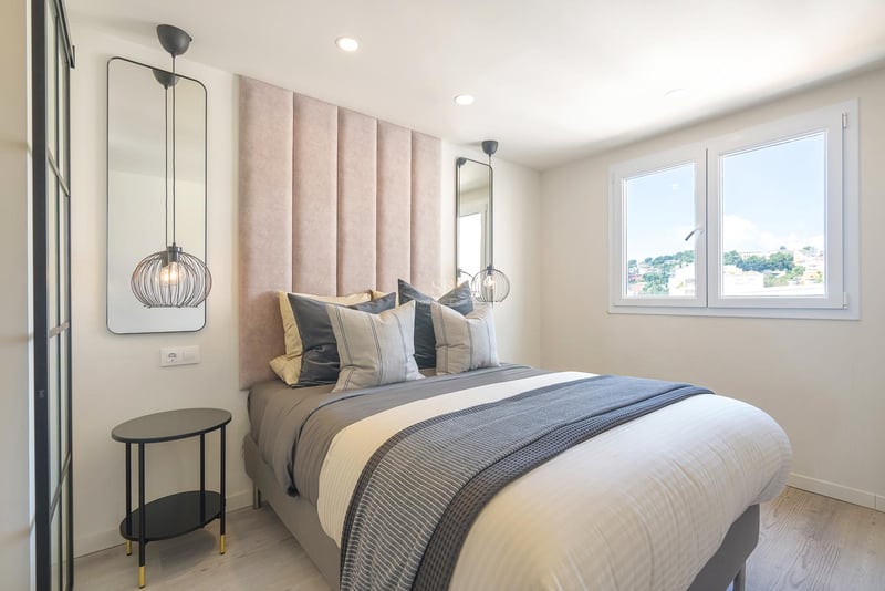 Chambre moderne avec tête de lit rose poudré, suspensions design, linge de lit gris et fenêtre lumineuse.