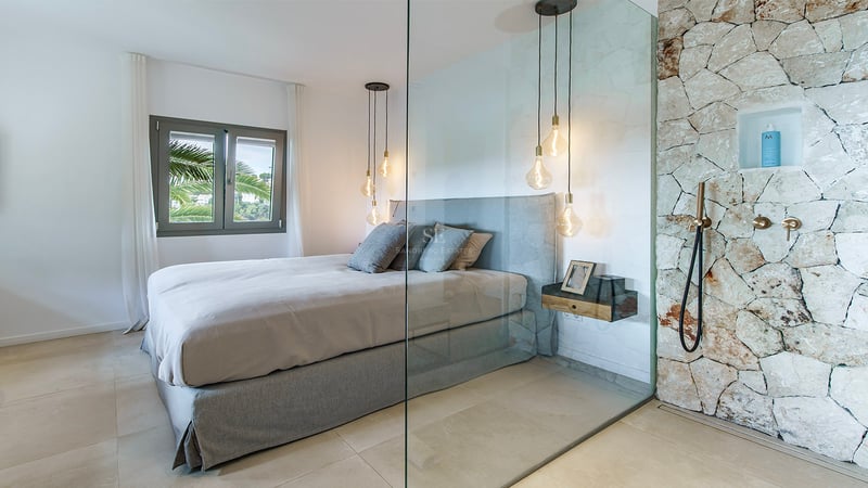 Chambre contemporaine avec lit gris, cloison en verre et douche en pierre rustique avec robinetterie en laiton.