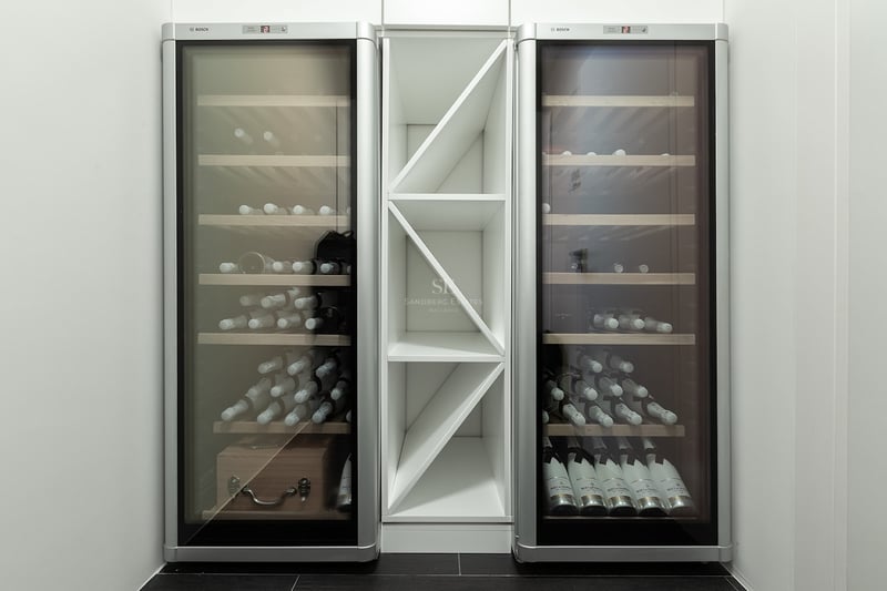 Deux élégantes caves à vin Bosch argentées encadrant une étagère à bouteilles blanche dans une pièce minimaliste.