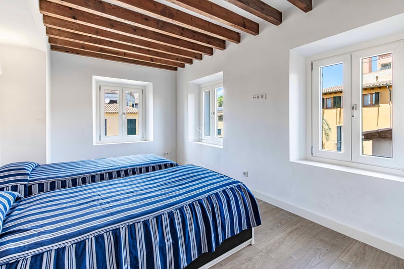 Chambre lumineuse avec deux lits simples, plafond à poutres en bois et fenêtres donnant sur la vieille ville.
