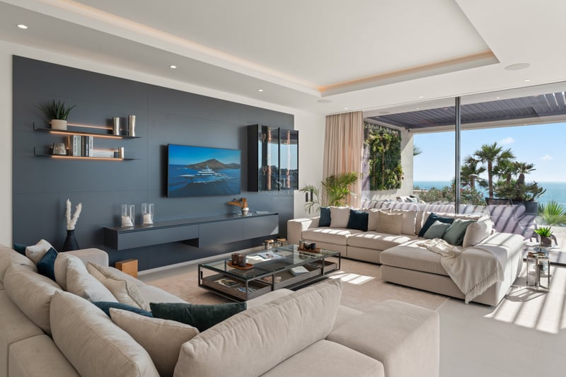 Salon lumineux avec canapés beiges, mur d'accent anthracite, télévision et baies vitrées ouvrant sur une terrasse vue mer.