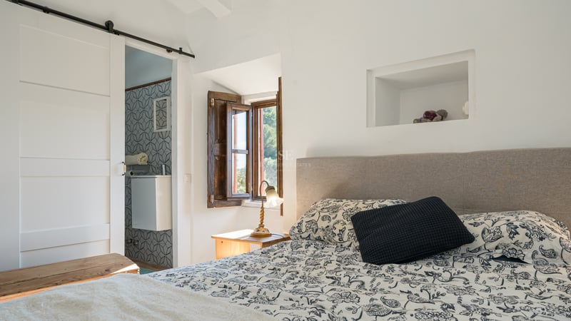 Chambre ensoleillée avec couvre-lit à motifs, volet en bois traditionnel et vue sur une salle de bain carrelée.