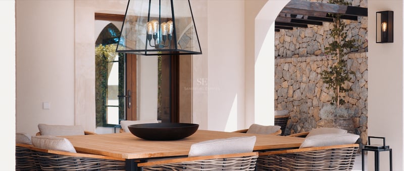 Table à manger en bois et chaises tressées sur une terrasse de luxe avec mur en pierre et lanterne design.