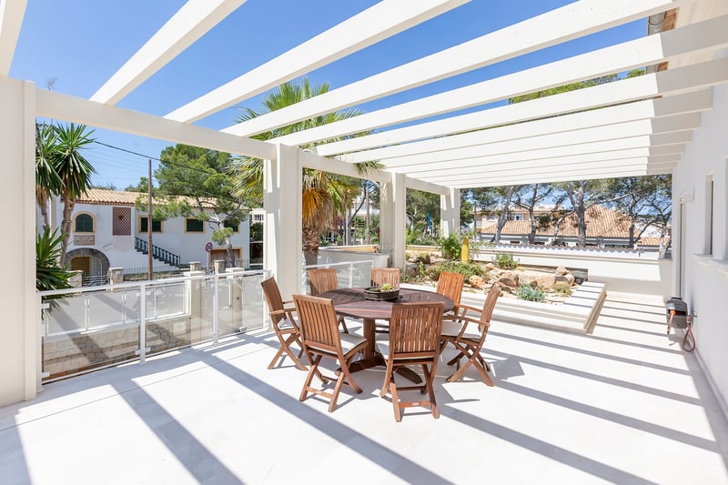 Terrasse ensoleillée avec une pergola blanche, une table ronde en bois et des chaises, entourée de palmiers.