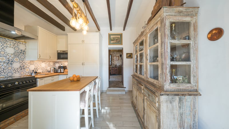 Cuisine lumineuse avec poutres de plafond en bois sombre, armoires blanches, îlot avec tabourets et grand buffet vintage.