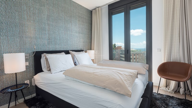 Chambre moderne avec mur tapissé bleu, lit blanc et grande fenêtre offrant une vue sur la mer et la ville.